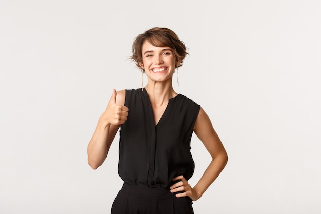 Attractive smiling woman showing thumbs-up in approval, recommending product, standing white.