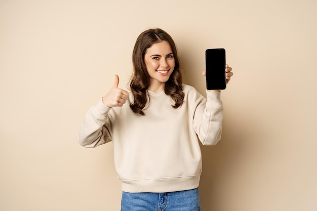 Attractive smiling woman showing thumbs up and smartphone screen standing over beige background