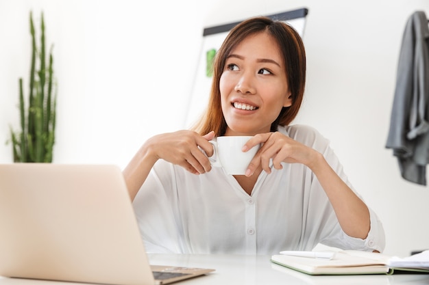 Attractive smiling young asian businesswoman working while sitting at the desk in office, holding cup of coffee