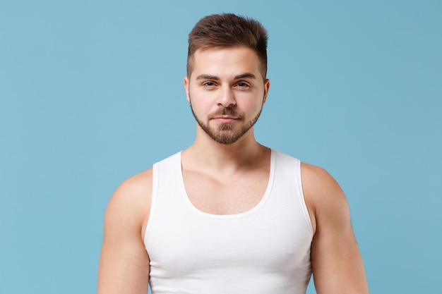 Attractive smiling young bearded guy 20s in white singlet posing isolated on pastel blue wall background studio portrait. Sport fitness healthy lifestyle concept. Mock up copy space. Looking aside.