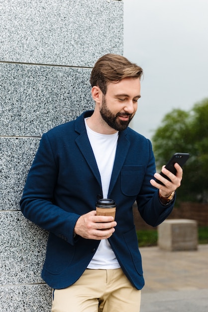 Attractive smiling young bearded man wearing jacket using mobile phone while standing outdoors at the city and drinking takeaway coffee