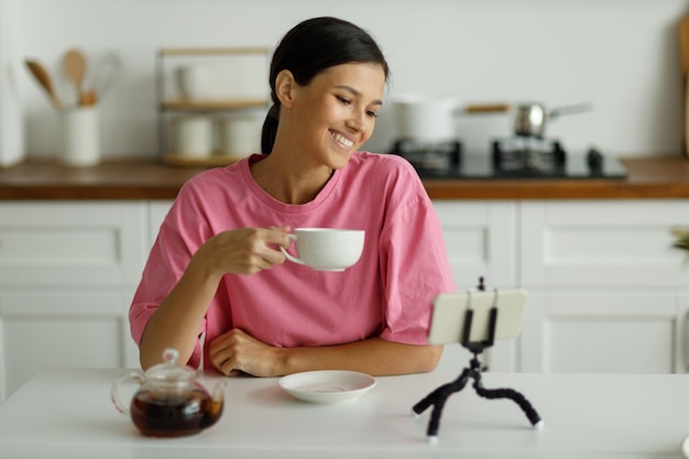 Attractive smiling young brunette woman in pink oversize Tshirt is drinking tea in kitchen Happy pretty girl with snowwhite smile greets friend communicating by video call online on mobile phone