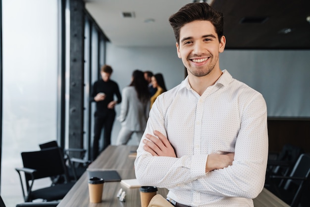 Attractive smiling young confident businessman in formal wear sitting in the office with his colleagues on a wall, arms folded