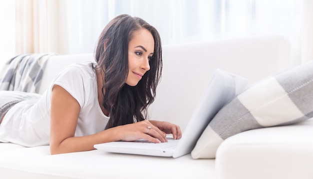 Attraente giovane donna sorridente in una t-shirt bianca e pantaloni della tuta sdraiata su un divano, scrivendo su un computer portatile appoggiato al cuscino.