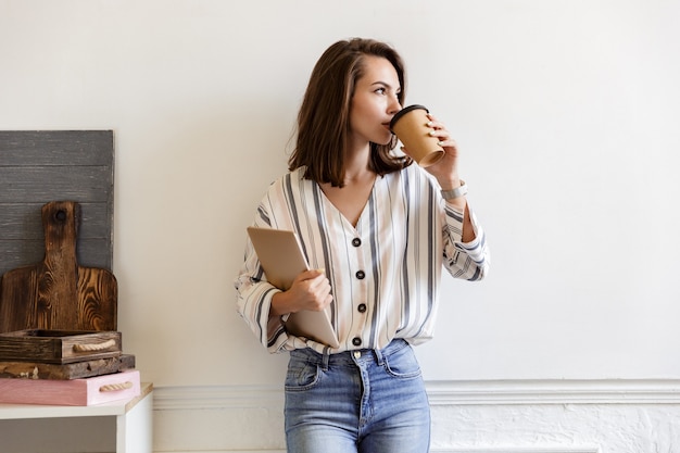 Attractive smiling young girl leaning on a wall while standing and holding laptop computer at home, drinking taleaway coffee