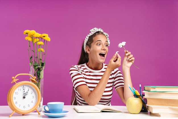 Attractive smiling young girl sitting at the desk isolated over pink wall, doing homework