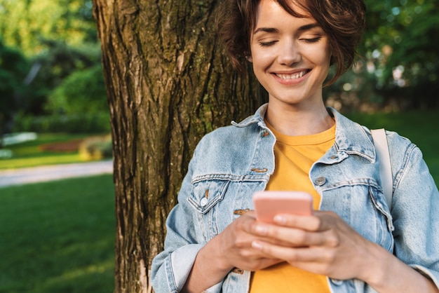 캐주얼 복장을 한 매력적인 미소 짓는 어린 소녀는 공원에서 야외에서 시간을 보내고, 나무에 기대고, 휴대 전화를 사용합니다.