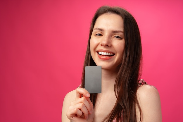 Привлекательная улыбающаяся молодая женщина, держащая черную кредитную карту на фоне розовой студии