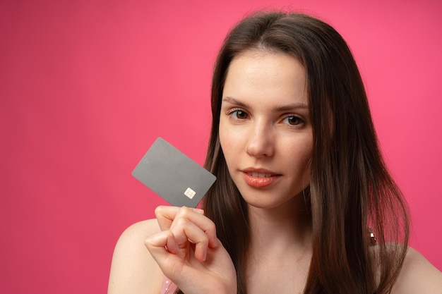 Attractive smiling young woman holding black credit card against pink studio background