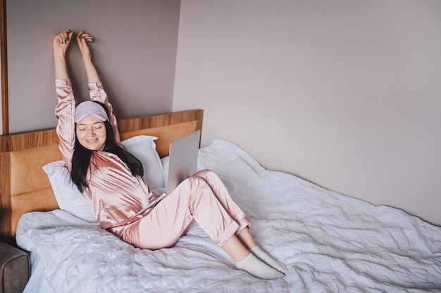 Attractive smiling young woman in pink silk pajamas and eye sleeping mask stretching in bed waking