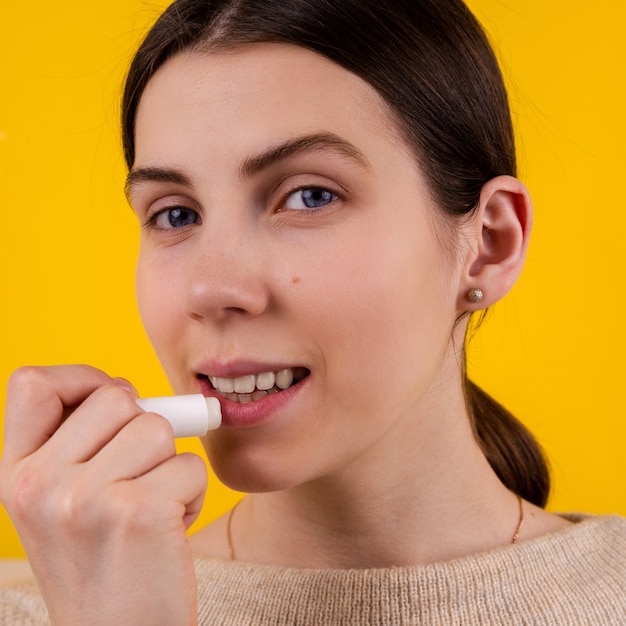 Attractive smiling young woman using hygienic lipstick on yellow background. lips care and protection.