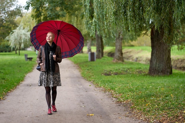 写真 巨大な赤い傘を持って秋の公園を歩いている魅力的な微笑む短い髪のブロンドの女性