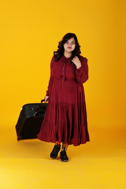 La donna attraente del viaggiatore del sud asiatico in vestito rosso intenso dell'abito ha posato allo studio su fondo giallo con la valigia