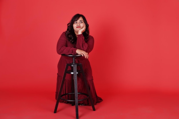 Attractive south asian woman in deep red gown dress posed at studio on pink background sitting on chair