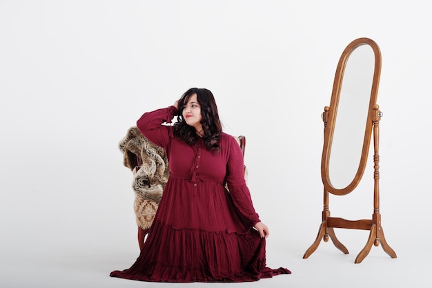 Attractive south asian woman in deep red gown dress posed at studio on white background against mirror and chair