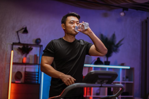 トレッドミルで実行されているフィットネス運動をしているボトルから水を飲む魅力的なスポーツ アジア人男性