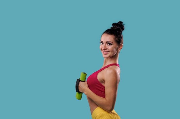 Attractive and sporty woman holds dumbbells and cute smiles to camera posing sideways on blue background