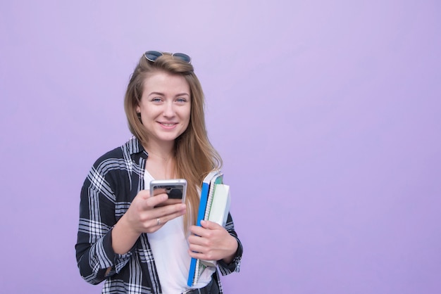 매력적인 학생 소녀 책, 노트북 및 스마트 폰 카메라를보고 웃 고 그녀의 손에 보라색 배경에 고립.