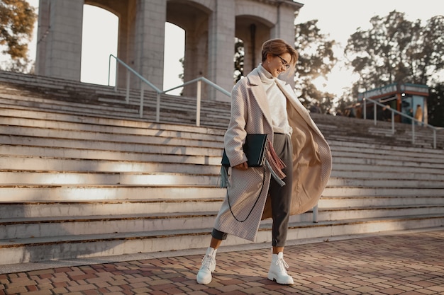 Attractive stylish fashionable woman walking in street in elegant style coat wearing glasses, bag and white boots