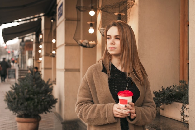 魅力的なスタイリッシュな女性は、ベージュの暖かいコートを着て、手にコーヒーを片手に路上に立ち、横を向きます。町の通りの春服の女の子