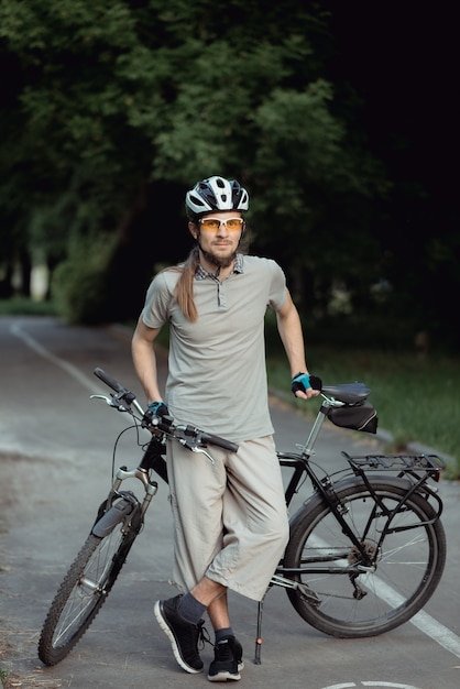 公園で自転車の横にポーズをとる魅力的なスタイリッシュな男。彼は自転車を持っています。