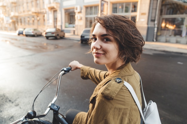 Attraente donna elegante che indossa cappotto in sella a una bicicletta su una strada cittadina