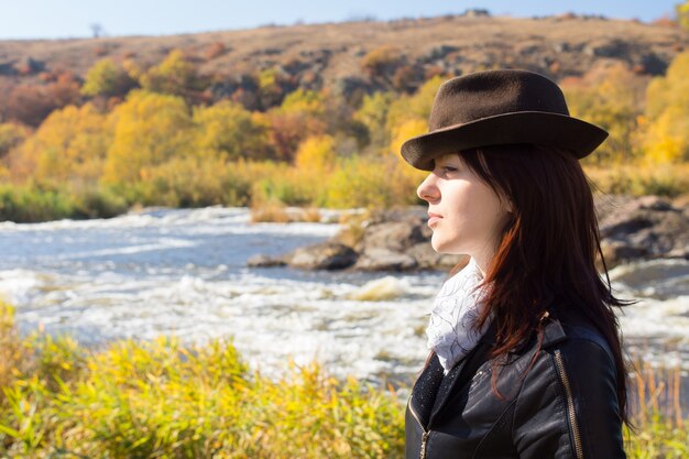 Attractive stylish woman with long brown hair wearing a trendy hat, scarf and jacket posing in countryside in front of a river, with copyspace, side view