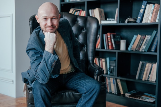 Attractive successful adult bald smiling man with beard in suit sitting in chair in his office