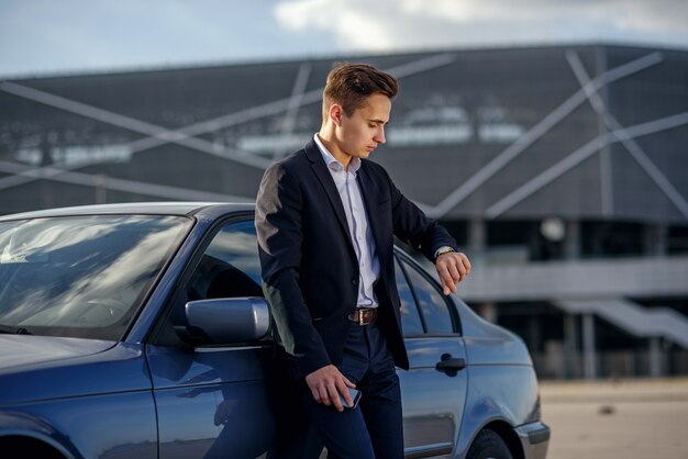 彼の車の近くの時計を探しているビジネススーツで魅力的な成功した青年実業家