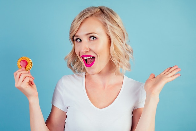 Attractive surprised and amazed woman blonde with make-up and lipstick on lips color fuchsia keeps a high-calorie cookie biscuit in hand in the studio on a blue background