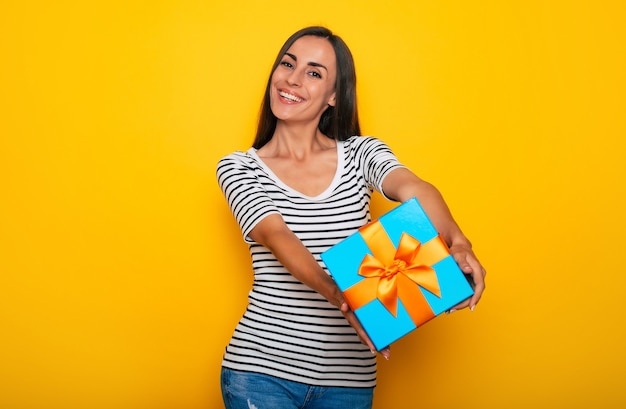 Attractive surprised excited young smiling woman is posing with big cool gift box in hands isolated on yellow background