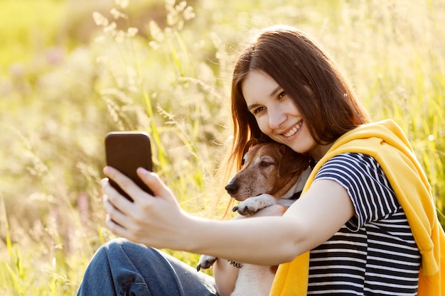 魅力的な十代の少女は、携帯電話のカメラを使用して自分と犬の写真を撮っています。