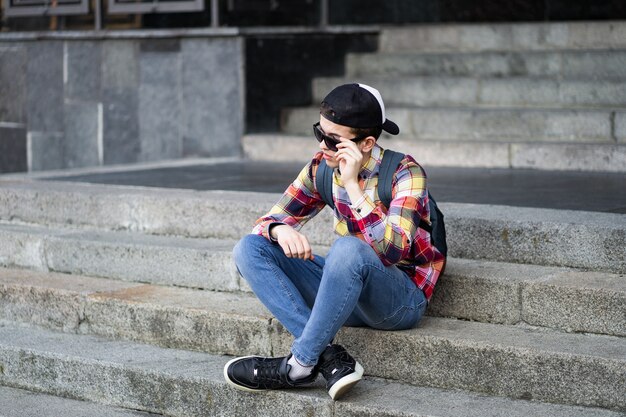 Attractive teenage boy in checkered shirt, blue jeans, sunglasses and baseball cap sitting on a college campus on the stairs waiting for someone or something. Education technology lifestyle. Fashion guy