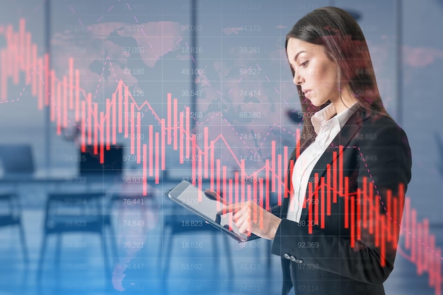 Attractive thoughtful european businesswoman using tablet with falling red business graph grid on blurry office interior background Crisis recession and stock market concept Double exposure