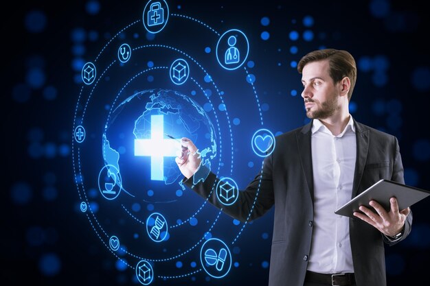 Attractive thoughtful young businessman with tablet using creative glowing blue medical hologram with cross and globe on dark background Science healthcare system and futuristic pharmacy concept