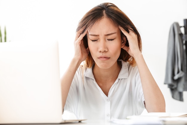 Attractive tired young asian businesswoman working while sitting at the desk in office, working on laptop computer, suffering from a headache