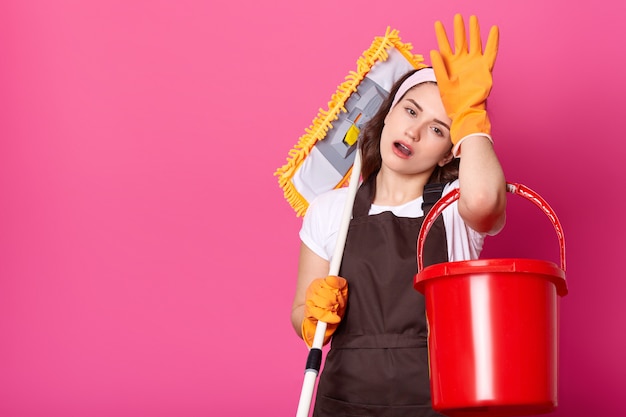 Attractive tired young female keeps hand on forehead, puts mop on shoulder.