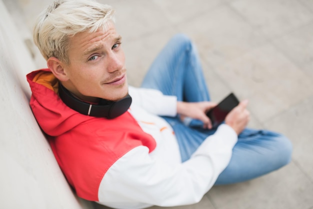 Photo attractive above view of young blond man looking up and enjoying weather in city park waiting for his friends texting message with headphones on the neck outside in the park