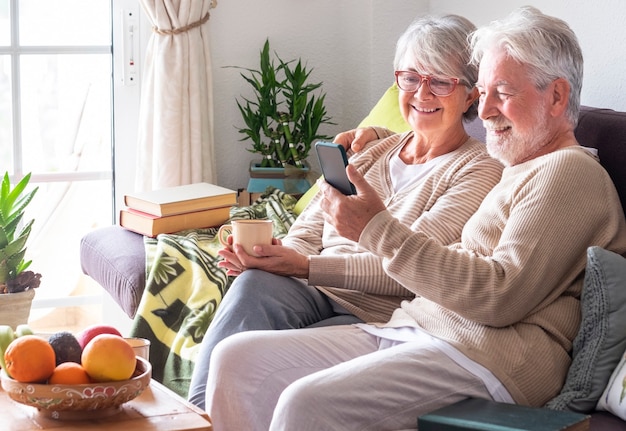 Привлекательные седые старшие пары отдыхают дома на диване, вместе глядя на смартфон во время видеозвонка с семьей или друзьями. улыбающиеся пожилые люди наслаждаются беспроводной технологией