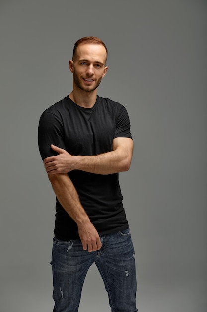 An attractive white man in jeans and black shirts tands in full growth against a gray background Copy space