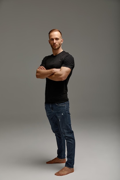 An attractive white man in jeans and black shirts tands in full growth against a gray background Copy space