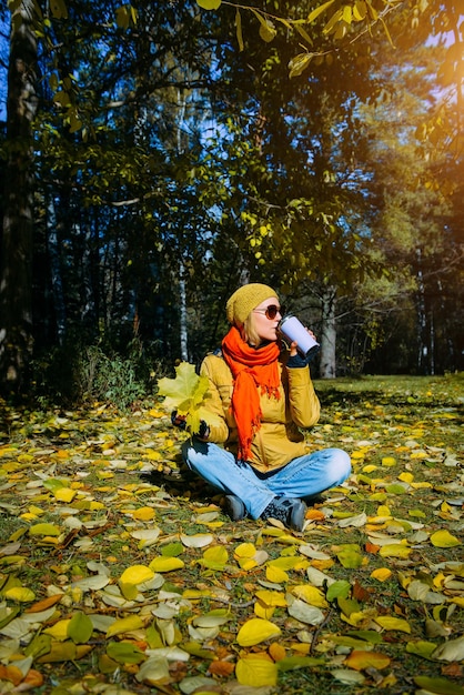 Attractive woman in autumn park sitting on yellow foliage in sunlight Pretty girl holding maple leaves and drinking coffee Happy healthy lifestyle