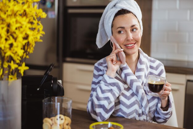 An attractive woman in bathrobe using smartphone while enjoying morning coffee at home.