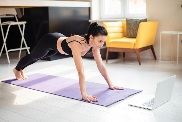 Attractive woman in black sport clothes training on yoga mat and watching tutorial on modern laptop Concept of domestic workout