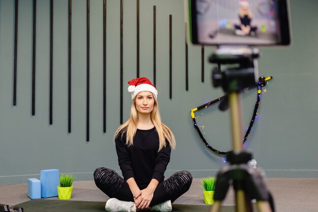 Attractive woman blogger at Santa hat in sportswear records exercises for workout for her vlog.