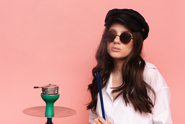 An attractive woman in a cap and glasses smokes a hookah, sheesha and enjoys smoking. Pink solid color background.