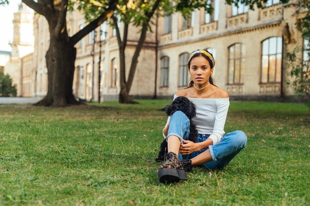 Attractive woman in casual clothes sitting on the grass in the park with a cute little dog