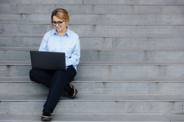 Attractive woman in casual clothing and eyewear enjoying fresh air while working in modern laptop. Pretty blonde using modern gadgets for work while sitting on steps.