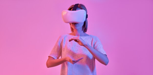 Attractive woman in casual wear using virtual reality glasses holding something in palms isolated on neon pink