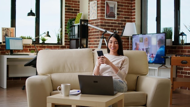 Donna attraente che chiacchiera sullo smartphone mentre si siede sul divano a casa e sorride di cuore alla telecamera. felice giovane adulto che invia messaggi sul moderno telefono cellulare mentre lavora in remoto.